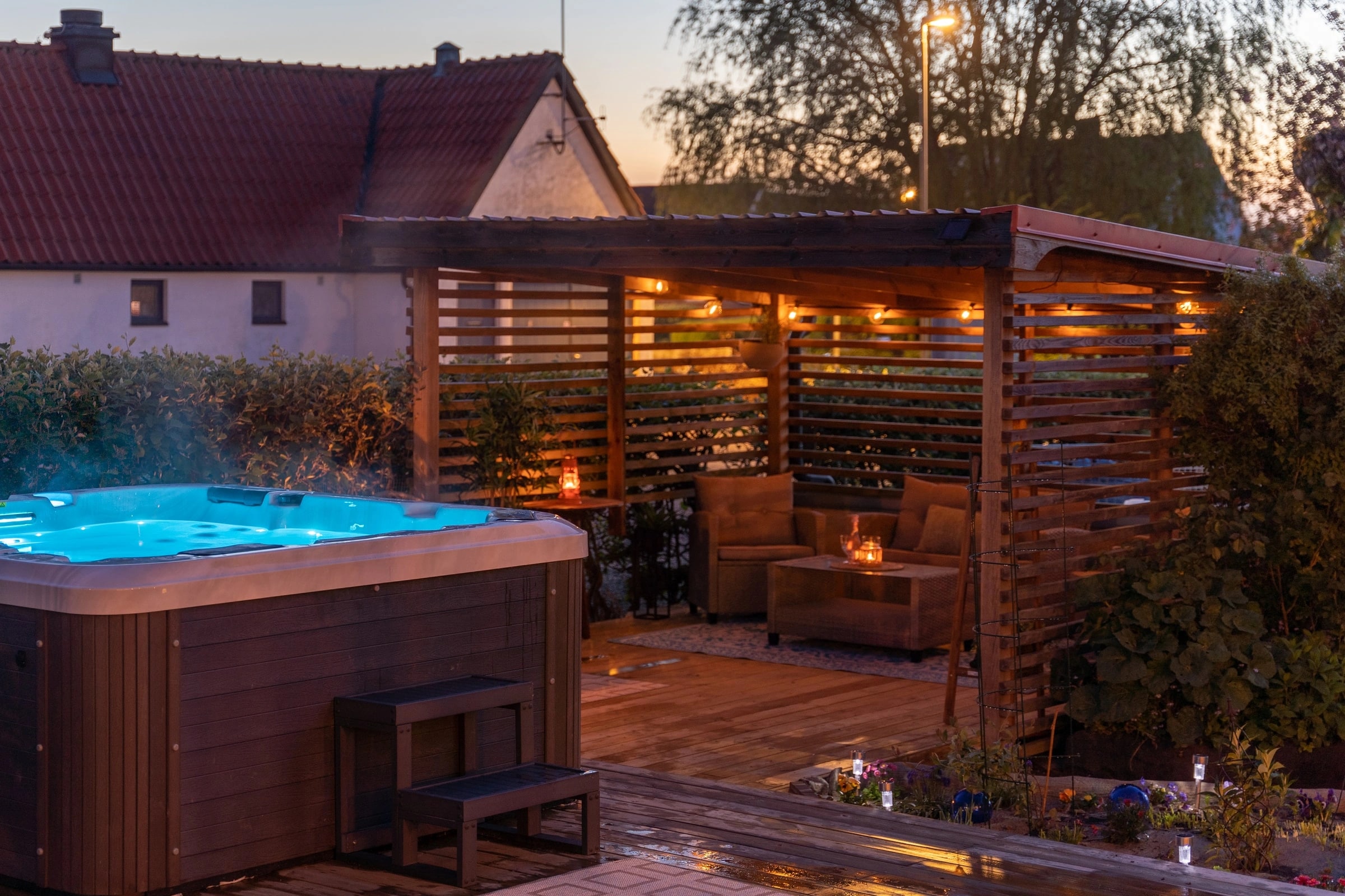 Uteplats med pergola och badtunna i skymningsljus, lyktor tända under taket