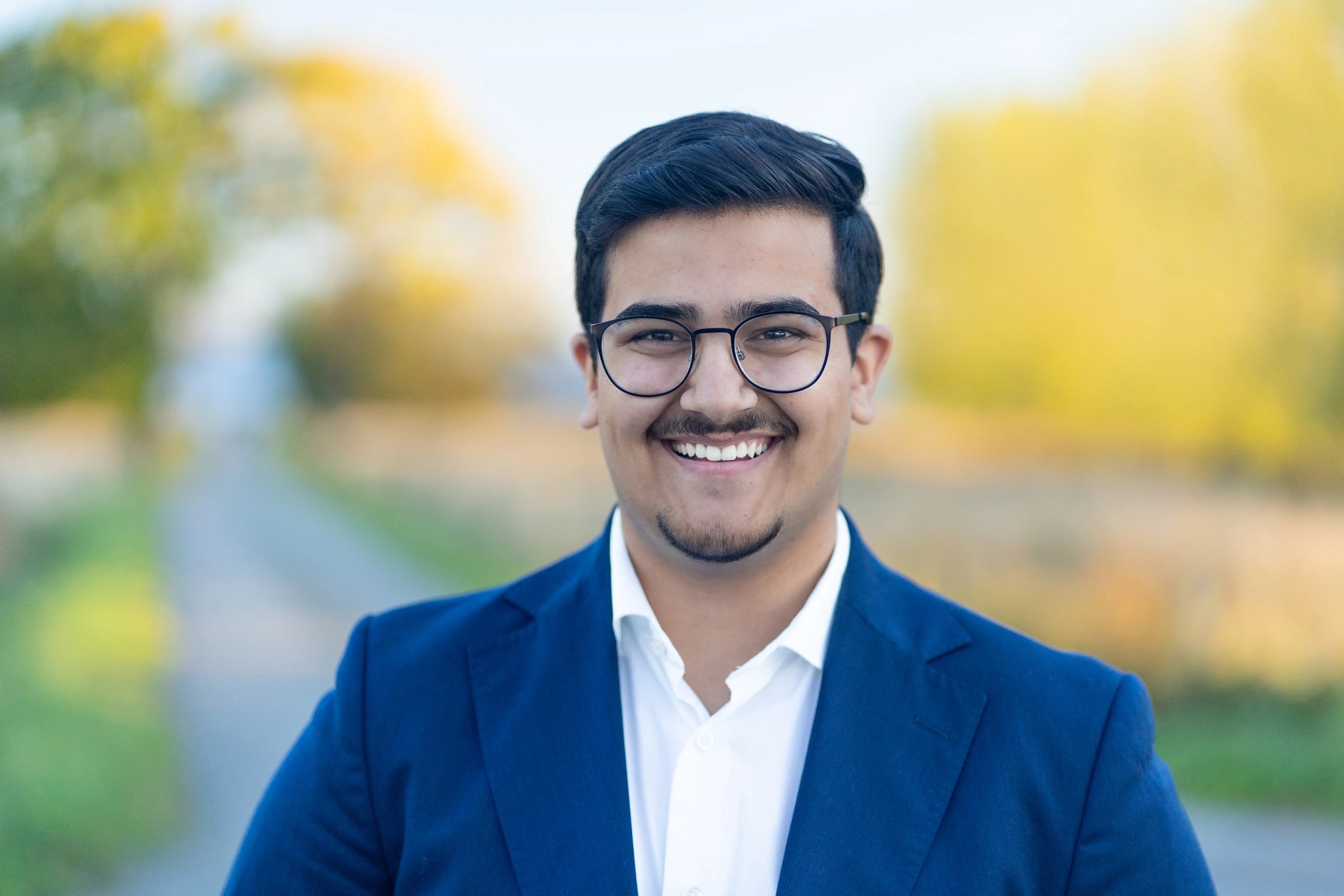Leende man i blå kavaj, fotograferad utomhus med en höstgul allé bakom sig
